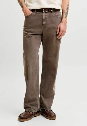 Homme portant un jean marron taille haute à jambe droite avec une ceinture noire et des chaussures en cuir marron. Tatouages visibles sur les bras.