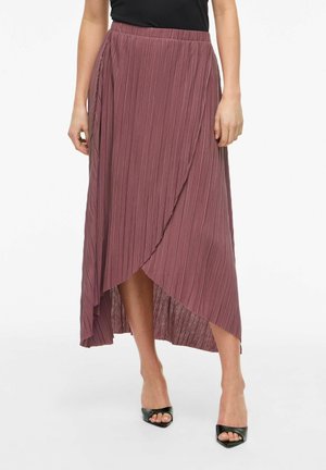 Kobieta nosząca plisowaną spódnicę w kolorze fioletowym o asymetrycznym, kopertowym przodzie, zestawioną z czarnymi, otwartymi szpilkami i czarnym topem.