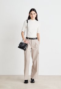 Una giovane donna sta in piedi con sicurezza, indossando un top aderente color crema, pantaloni beige a vita alta e tenendo una borsa nera con una tracolla a catena.