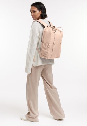 Růžový látkový batoh s polstrovanými ramenními popruhy, vrchním madlem na nošení, několika kapsami a zjednodušeným obdélníkovým tvarem.