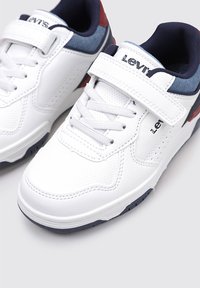 Zapatillas blancas con una parte superior sintética texturizada, acentos en azul marino y rojo en contraste, cierre de cordones y correa, y una suela de goma.