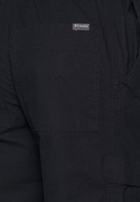 Svarta cargo-byxor med en strukturerad tyg, med en fackficka och en Columbia-logotagg. Designen visar sömnadsdetaljer.