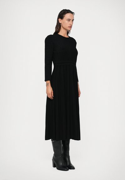 Vestido preto com mangas longas, textura canelada no corpete, saia fluida e botas de couro negras até o joelho com um salto robusto.