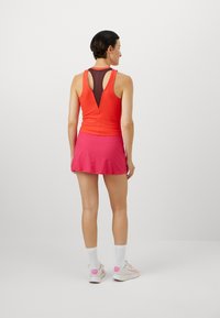 Gestreiftes rotes Tanktop mit einem dunklen Netzrücken und einem pinken, schwingenden Rock, kombiniert mit weißen Socken und Sneakers mit pinken Akzenten.