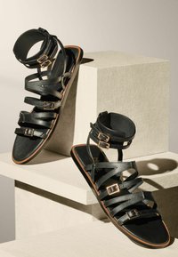 Mustade nahast sandaalid mitme rihmaga, ankle-buckle ja lamedal tallal. Sile tekstuur kuldsete metalldetailidega.
