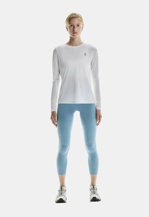 Vit långärmad träningströja med liten logotyp, matchad med ljusblå leggings. Modellen har på sig grå och vita löparskor. Enkel design.