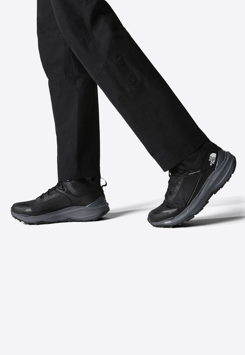 Czarne buty sportowe z teksturowanym wykończeniem, amortyzowaną podeszwą i odblaskowym detalem logo, w połączeniu z czarnymi spodniami.