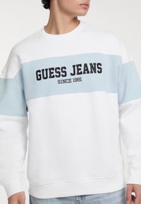 Felpa bianca con striscia orizzontale blu chiaro. Presenta il logo "GUESS JEANS" in nero, polsini a costine e una texture morbida.
