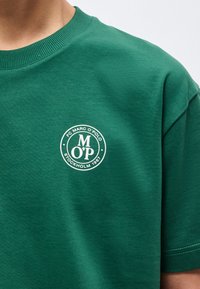 Πράσινο t-shirt με λαιμόκοψη τύπου crewneck και λευκό κυκλικό λογότυπο που γράφει "FC Marc O'Polo Stockholm 1967" στο αριστερό στήθος.