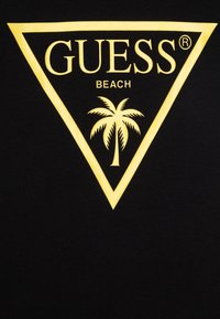 T-shirt noir avec un logo triangulaire jaune portant le texte "GUESS BEACH" et une illustration de palmier à l'intérieur du triangle.