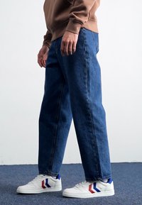 Dunkelblaue Jeans aus Denim, lässig geschnitten, mit Seitentaschen und geradem Bein. Kombiniert mit weißen Turnschuhen mit roten und blauen Akzenten.