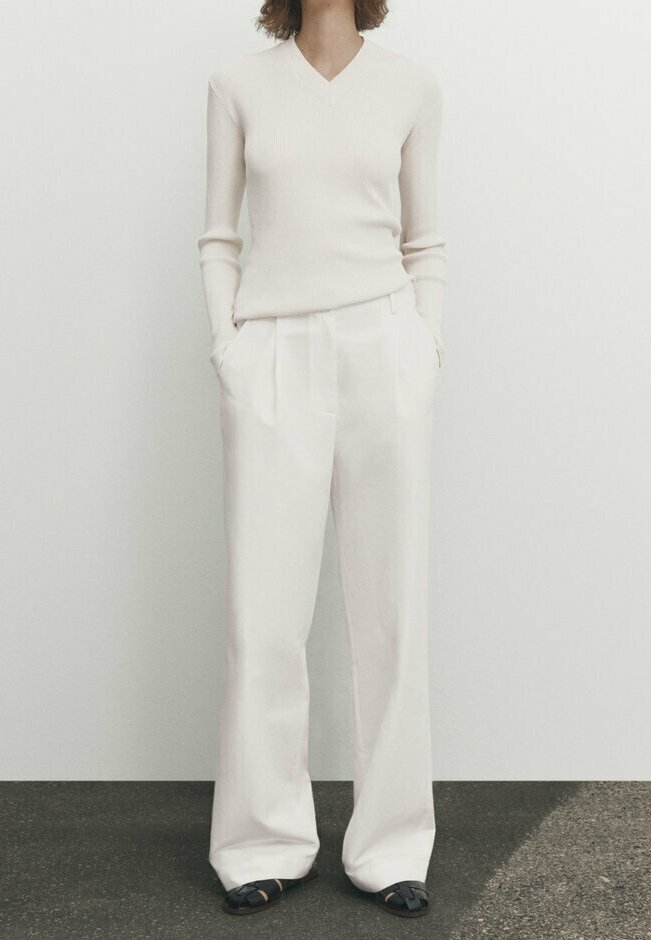 Personne portant un haut à manches longues côtelé crème, un pantalon blanc taille haute à jambe large et des chaussures noires, debout les mains dans les poches.