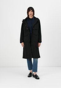 Casaco parka preto com capuz, fecho éclair, dois bolsos frontais e um corte relaxado. Usado sobre uma camisa azul escura e jeans azul claro.