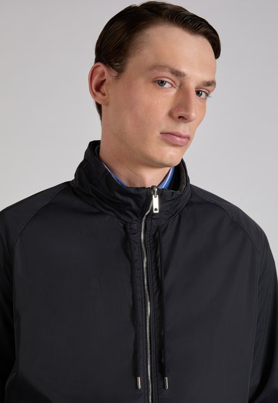 Młody mężczyzna z zaczesanymi do tyłu włosami, ubrany w czarną kurtkę na zamek nałożoną na niebieską koszulę, patrzący w bok na jednolitym jasnym tle.