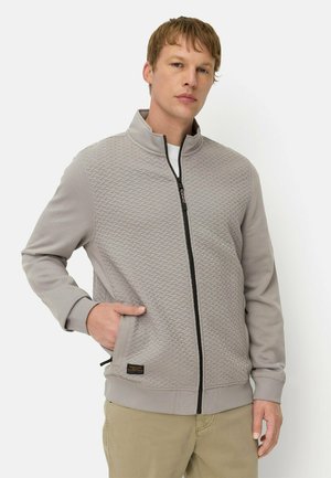 Jonge man draagt een lichtgrijs gestructureerd jack met rits en hoge kraag en beige broek, staand met één hand in de zak.
