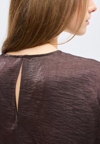 Γυναίκα με μακριά καστανά μαλλιά που φοράει ένα γυαλιστερό σκούρο καφέ τοπ με μια μικρή κατακόρυφη άνοιγμα στο πίσω μέρος κοντά στη λαιμόκοψη.