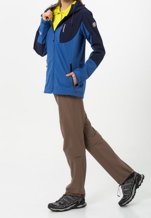 Persoon draagt een blauw en marineblauw jack, gele shirt, bruine broek en zwarte wandelschoenen, loopt met één hand in de zak van het jack.