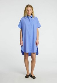 Abito camicia azzurro chiaro con maniche corte, caratterizzato da un orlo asimmetrico, una texture morbida e sottili dettagli di bottoni rosa.