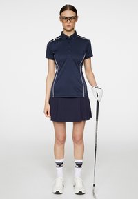 Marinblå polo med vita detaljer, tillsammans med en marinblå plisserad kjol. Tillbehören inkluderar vitt golfhandske och sportiga solglasögon.