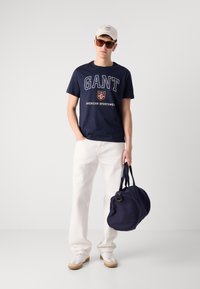 Tricou din bumbac bleumarin cu logo "GANT", șapcă crem, pantalonii albi și un rucsac bleumarin. Modelul poartă ochelari de soare și adidași albi.