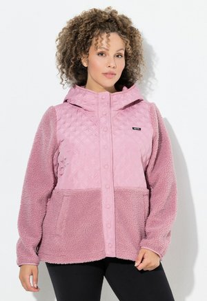 Vrouw met krullend haar die een roze doorgestikte en fleecetrui jas met capuchon en drukknopen draagt, staand tegen een effen witte achtergrond.