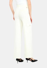Pantaloni bianchi a gamba larga con vita aderente, tasca laterale cargo e rifiniture in tono oro, abbinati a scarpe con tacco nere.