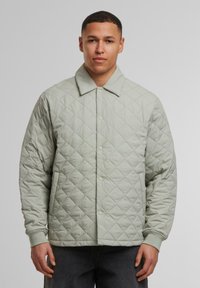 Grüne gesteppte Jacke mit einer Knopfleiste, großem Kragen, strukturiertem Stoff und gerippten Bündchen, getragen über dunklen Jeans.