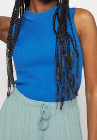 Sininen ribbikuvioinen hihaton toppi, jossa on teksturoitu chevron-kuvio, yhdistettynä vaaleanvihreään naruvyötäröskorttiin.