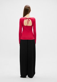 Rdeča dolga majica s krožnim izrezom na hrbtu, zvezana z trakcem; v kombinaciji s črnimi širokimi hlačami; gladka tekstura.