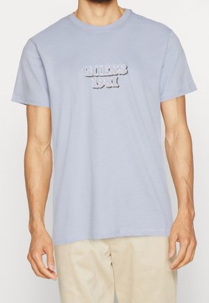 Homme portant une chemise à manches courtes bleu clair avec « GUESS 1981 » brodé sur la poitrine et un pantalon beige sur fond blanc.