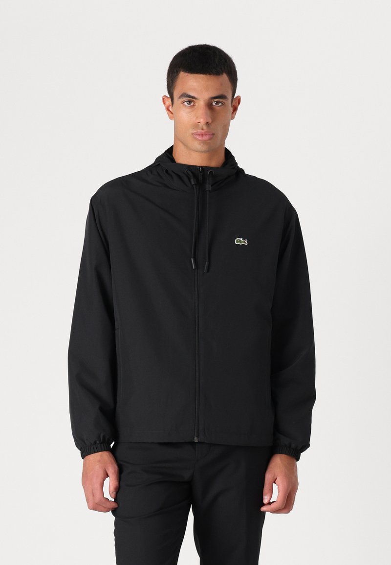 Veste noire avec capuche, poignets élastiques et fermeture éclair à l'avant. Dotée d'un petit logo Lacoste vert sur la poitrine gauche. Texture de tissu lisse.