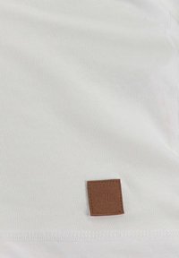 T-shirt bianco realizzato in morbido cotone. Presenta un quadrato in pelle marrone con testo impresso vicino all'orlo e rifinito con un filo di contrasto.