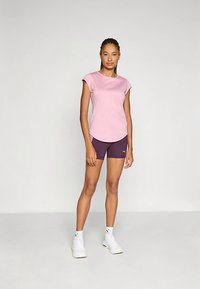 Rožinės spalvos trumpomis rankovėmis sportinis marškinėlis derinamas su tamsiai violetiniais šortais. Modelis stovi ant neutralaus fono, dėvi baltus sportinius batelius.