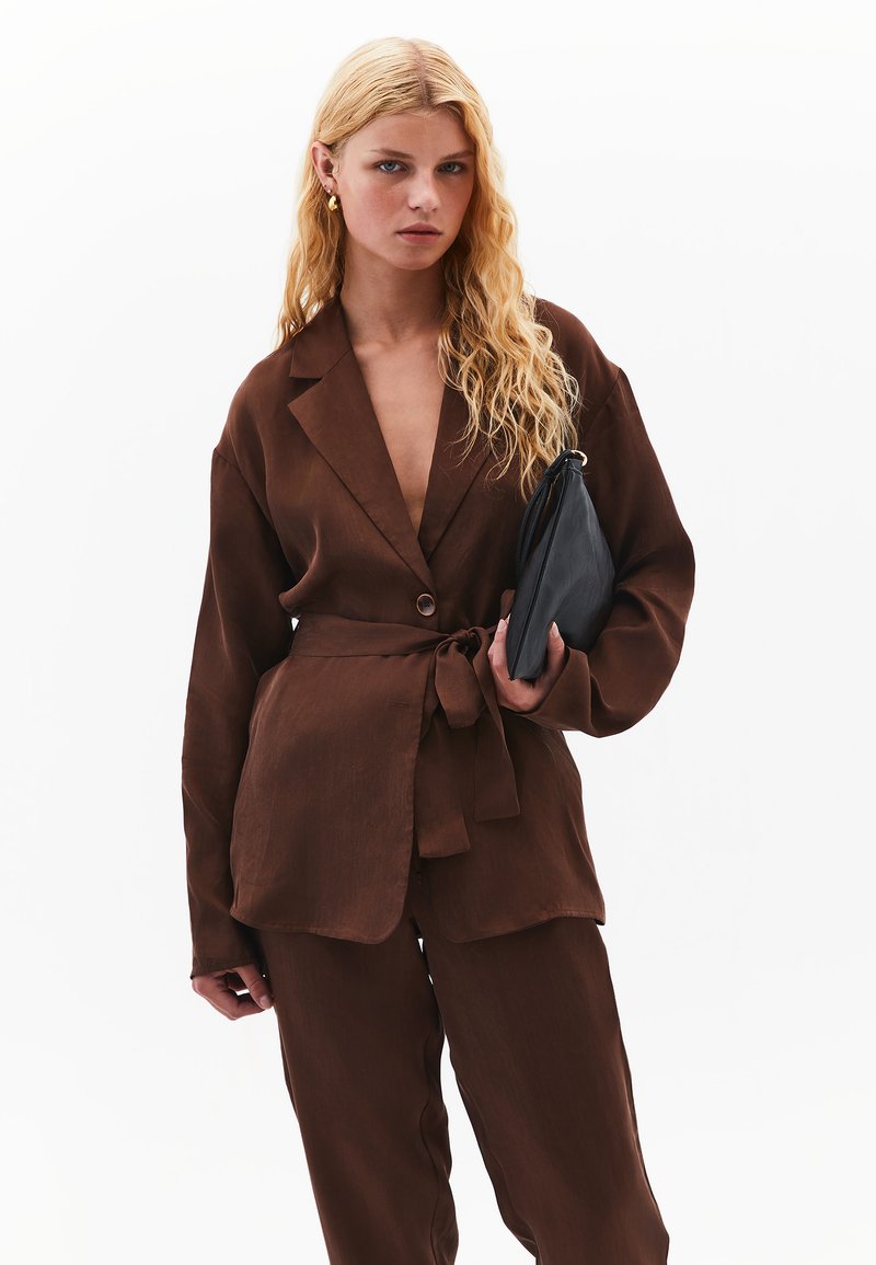 Hnědý, lehký, přizpůsobený blejzr s hlubokým výstřihem do V a páskem; doplněný širokými kalhotami ve stejném odstínu. Doplňky zahrnují černou kabelku.