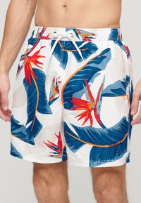 Bañadores para hombre con un fondo blanco y hojas tropicales en azul y verde, además de patrones florales rojos. Incluyen una cintura ajustable con cordón.