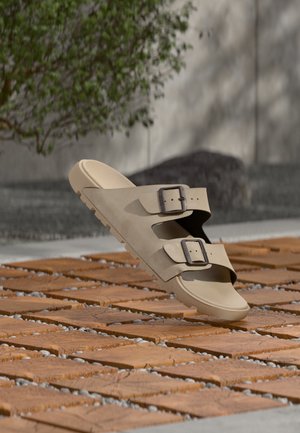 Beige sandal med to justerbare spænder, der svæver over træfliser udendørs med en grøn plante og en grå væg i baggrunden.