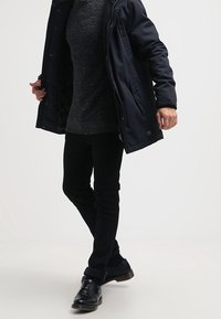 Long manteau noir avec capuche, associé à un pull gris foncé et un pantalon noir. Comprend un tissu texturé et des chaussures habillées noires.