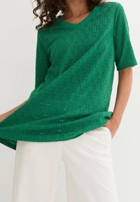 Blusa verde a maniche corte con scollatura a V realizzata in tessuto testurizzato con motivi aperti. Abbinata a pantaloni bianchi con una vestibilità comoda.