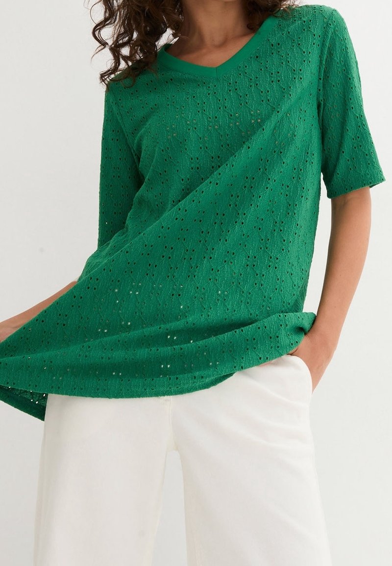 Blusa verde a maniche corte con scollatura a V realizzata in tessuto testurizzato con motivi aperti. Abbinata a pantaloni bianchi con una vestibilità comoda.