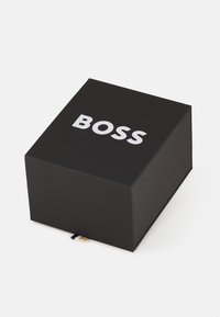 Musta ristkülikukujuline kast, millel on mati viimistlusega pind ja mille peal on julge valge "BOSS" logo. Ühel küljel on nähtav väike kangast silt.