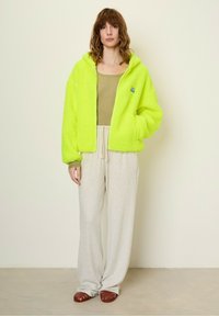 Neon green, fluffy fleece jacket with zipper and hood; worn over a light olive tank and paired with off-white sweatpants and brown sandals.