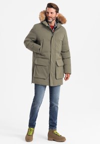 Olivgrüne Parka mit gefütterter Kapuze aus Fell, mehreren vorderen Taschen, Reißverschluss und gestepptem Muster. Getragen mit blauen Jeans und braunen Stiefeln.