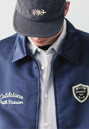 Person trägt eine marineblaue Jacke mit der Aufschrift „Coldstone Path Division“ und einem Sportemblem, ein gestreiftes Hemd mit Knöpfen und eine dunkle Kappe mit Stickerei von Vogel und Blumen.