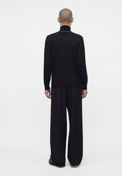 Personne à la tête rasée portant un pull à col roulé noir et un pantalon noir à jambes larges, debout de dos devant un fond uni.