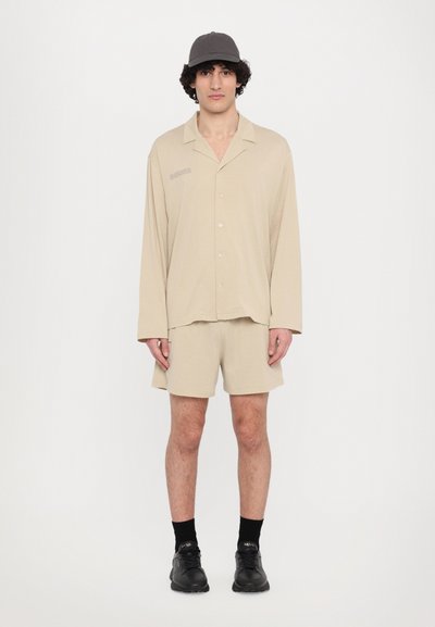 Beige Langarmhemd mit Knöpfen und lockerem Schnitt, kombiniert mit passenden Shorts. Das Model trägt eine dunkelgraue Kappe und schwarze Schuhe.