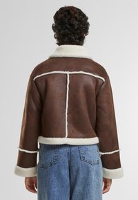Giacca in shearling marrone con rifiniture crema, design corto e maniche ampie. Le cuciture notevoli creano un motivo geometrico sulla schiena.