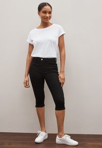 Femme debout contre un mur uni, portant un haut blanc à manches courtes, un pantalon noir court et des baskets blanches, souriant légèrement.