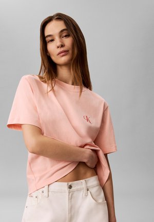 Ung kvinde med langt brunt hår iført en lyserød kortærmet skjorte og hvide bukser, der holder sin skjorte nær taljen.