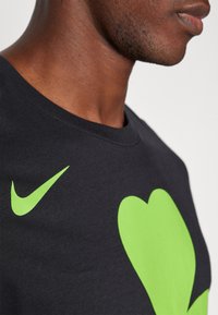Camiseta de algodón negra con un gran gráfico de corazón verde y un pequeño logotipo de Nike en verde. Diseño de cuello redondo con una textura suave.