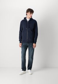 Felpa blu navy con zip e cappuccio regolabile, tessuto testurizzato, dettaglio del logo sul davanti, abbinata a jeans scuri e sneakers bianche.